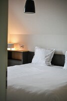 Uma cama bem arrumada com lençóis brancos, uma mesa de cabeceira com abajur e uma cabeceira escura.