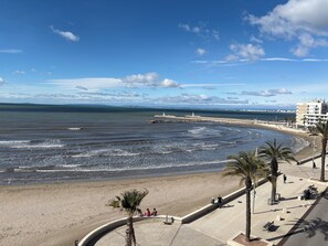 Vista costeira com praia arenosa, calçadão com palmeiras e um farol à distância.