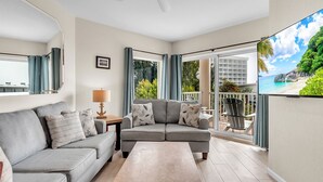 En moderne stue med en grå sofa, et sofabord og en loftsvifte. Der er en balkon med udsigt over en strand og grønt område.