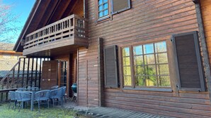 A wooden cabin with a balcony, large windows, and a dining area with chairs.