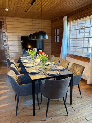 Een eetruimte met een houten tafel voor zes personen, omringd door stoelen met kussens, een televisie en een raam met gordijnen.