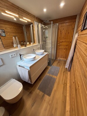 Een badkamer met houten wanden, een douchecabine van glas, twee wastafels en een toilet.
