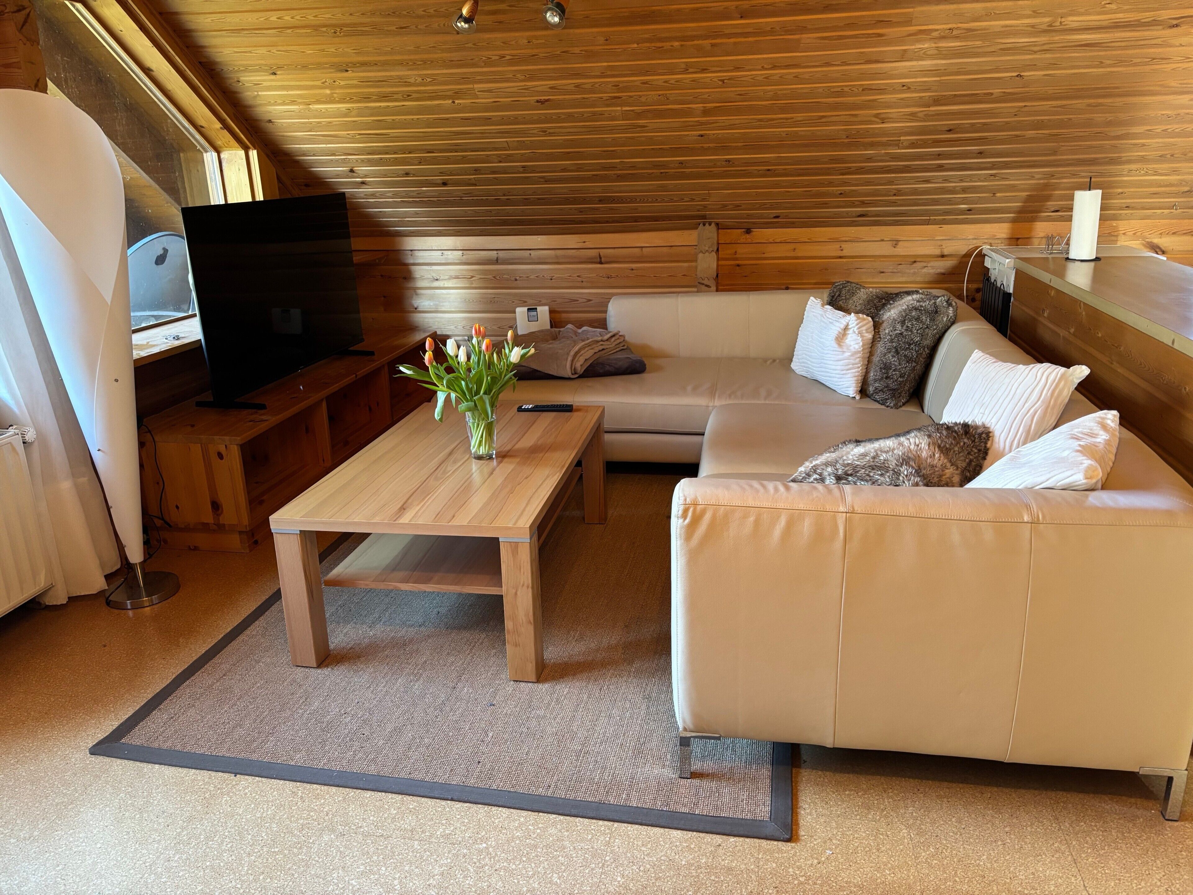 Ein gemütliches Wohnzimmer mit Holzbalkendecke, einem beigefarbenen Sofa, einem Holztisch, einem Fernseher und einer Vase mit Blumen.