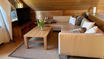 Ein gemütliches Wohnzimmer mit Holzbalkendecke, einem beigefarbenen Sofa, einem Holztisch, einem Fernseher und einer Vase mit Blumen.