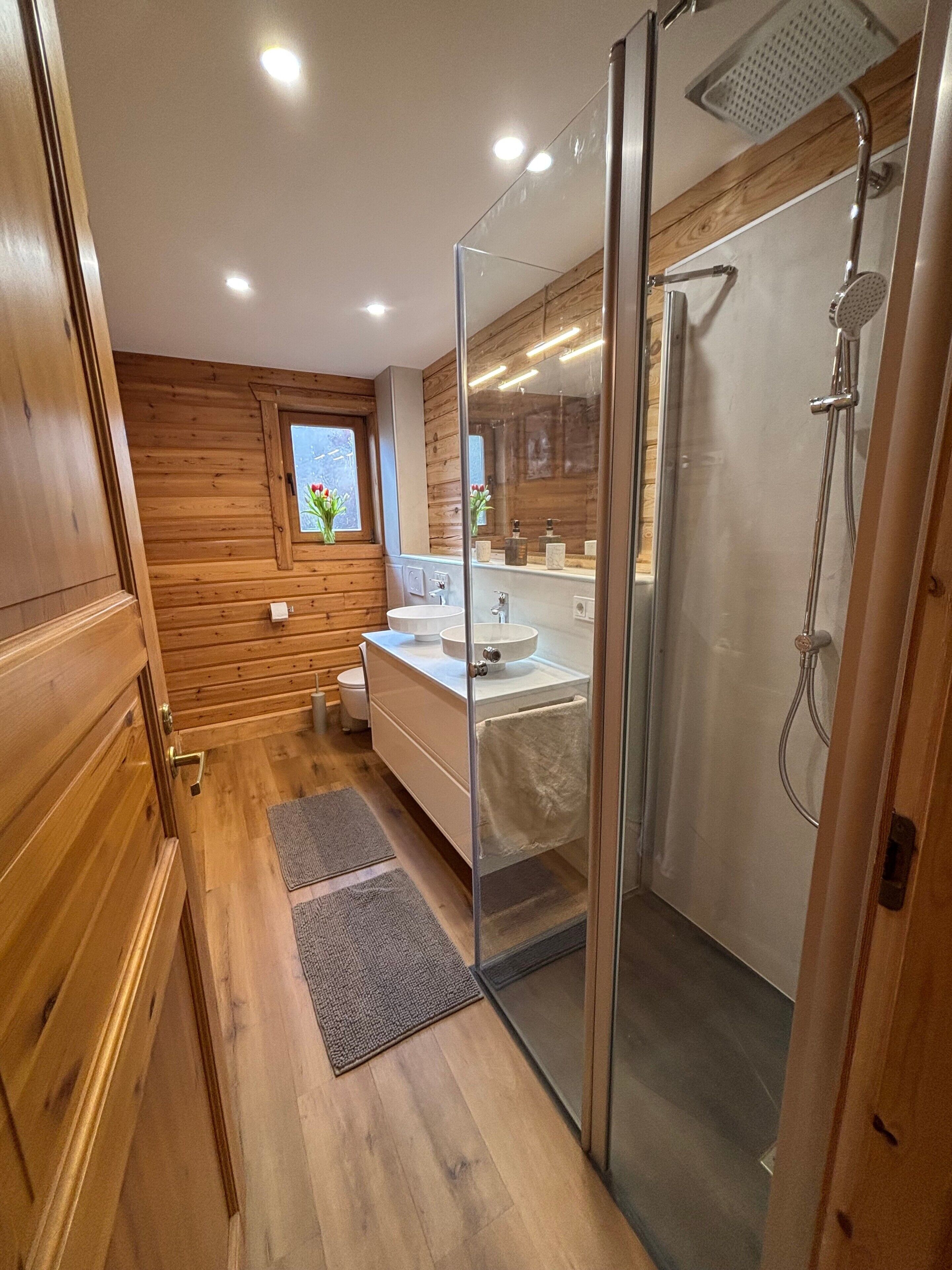 Ein modernes Badezimmer mit Holzvertäfelung, einer Glasdusche und einem Fenster mit Blumen.