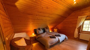 A cozy attic bedroom with a wooden sloped ceiling, a single bed with pillows and a blanket, a wicker chair, and a nightstand with a lamp.