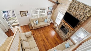 A spacious living area with a stone fireplace, wooden flooring, and a large TV mounted on the wall. - 0.2 Mi to Beach • 3 Ensuite Baths • Sleeps 10 • Walk to King & Prince (St Simons)