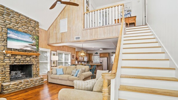 A spacious living room with a stone fireplace, a wooden staircase, and a kitchen in the background. - 0.2 Mi to Beach • 3 Ensuite Baths • Sleeps 10 • Walk to King & Prince (St Simons)