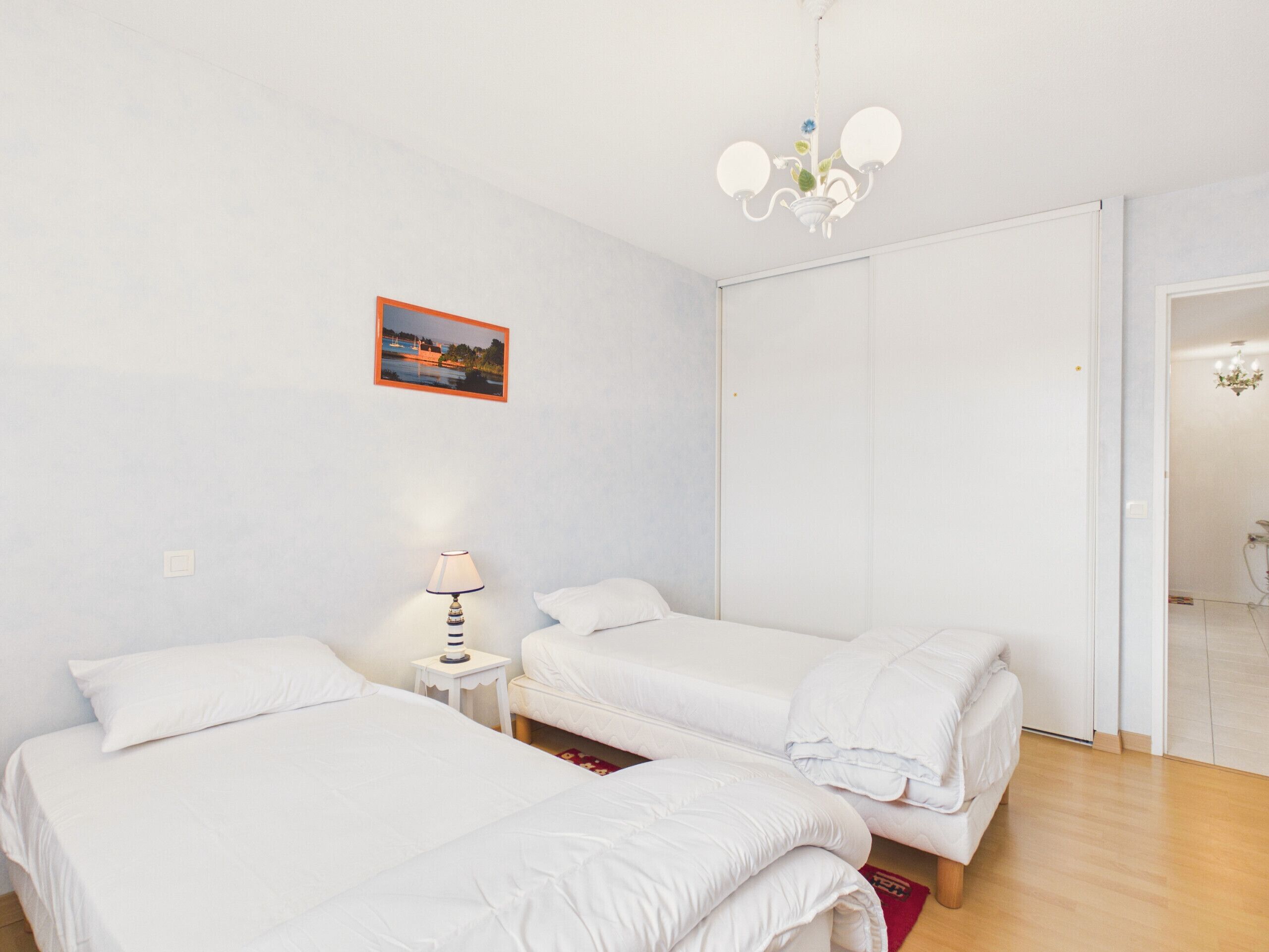 Ein Zimmer mit zwei Betten, einem kleinen Nachttisch mit Lampe, einem Bild an der Wand und einer Tür zu einem anderen Raum.