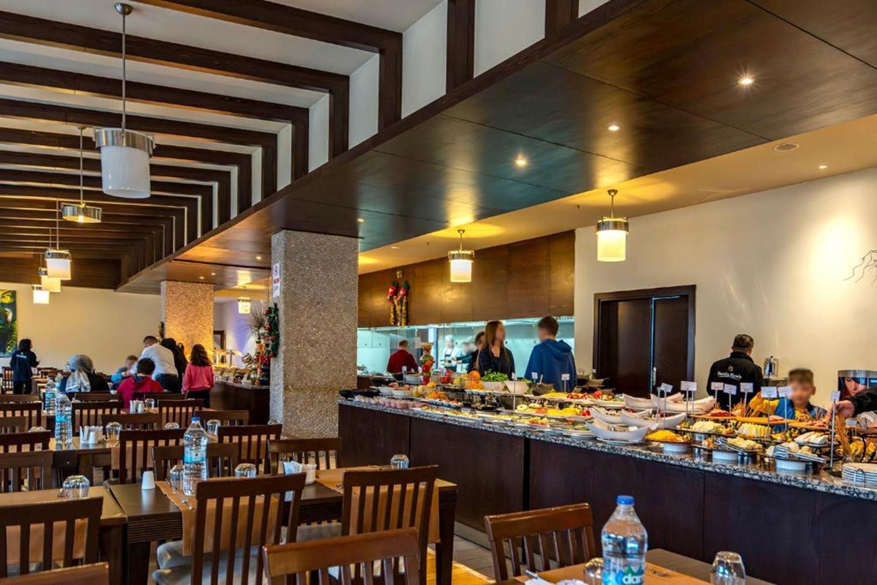 A buffet area with multiple food stations, wooden tables and chairs, and a kitchen in the background.