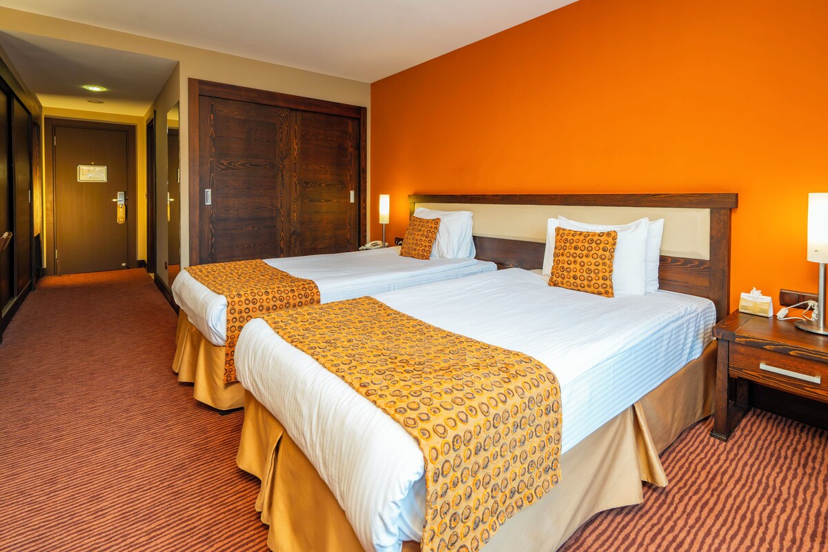 A hotel room with two beds, a wooden headboard, a carpeted floor, and a door in the background.