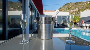 A table with two champagne flutes and a metal bucket with a bottle of champagne. The setting includes a poolside view with a red building and a hillside in the background. - Oceanfront Villa with Private Pool - Breathtaking Sunsets in Madeira (Tabua)