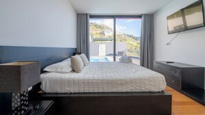 A modern bedroom with a large bed, a TV mounted on the wall, and a view of a pool and hills outside. - Oceanfront Villa with Private Pool - Breathtaking Sunsets in Madeira (Tabua)