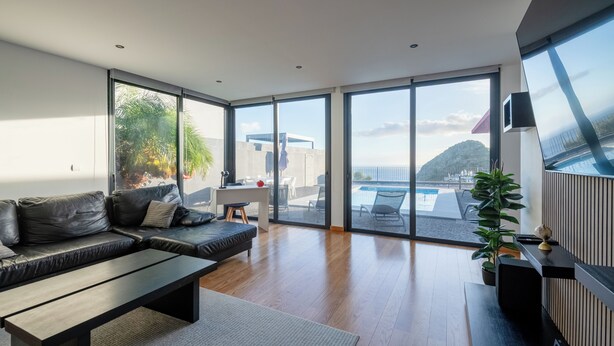 A modern living room with a sofa, coffee table, and large windows offering a view of a swimming pool and mountains. - Oceanfront Villa with Private Pool - Breathtaking Sunsets in Madeira (Tabua)