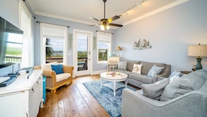 A spacious living room with a ceiling fan, a grey sofa, a wicker chair, a coffee table, and a view through the glass doors to the outdoors. - Lagoon-front Duplex w/Fishing Pier Sleeps 24/BM (Gulf Shores)