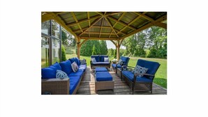 A covered outdoor seating area with wicker furniture and blue cushions, overlooking a lush garden. - Riverfront Estate in Paris, 5 luxury Suites indoor pool hot tub self serve spa (Brant)