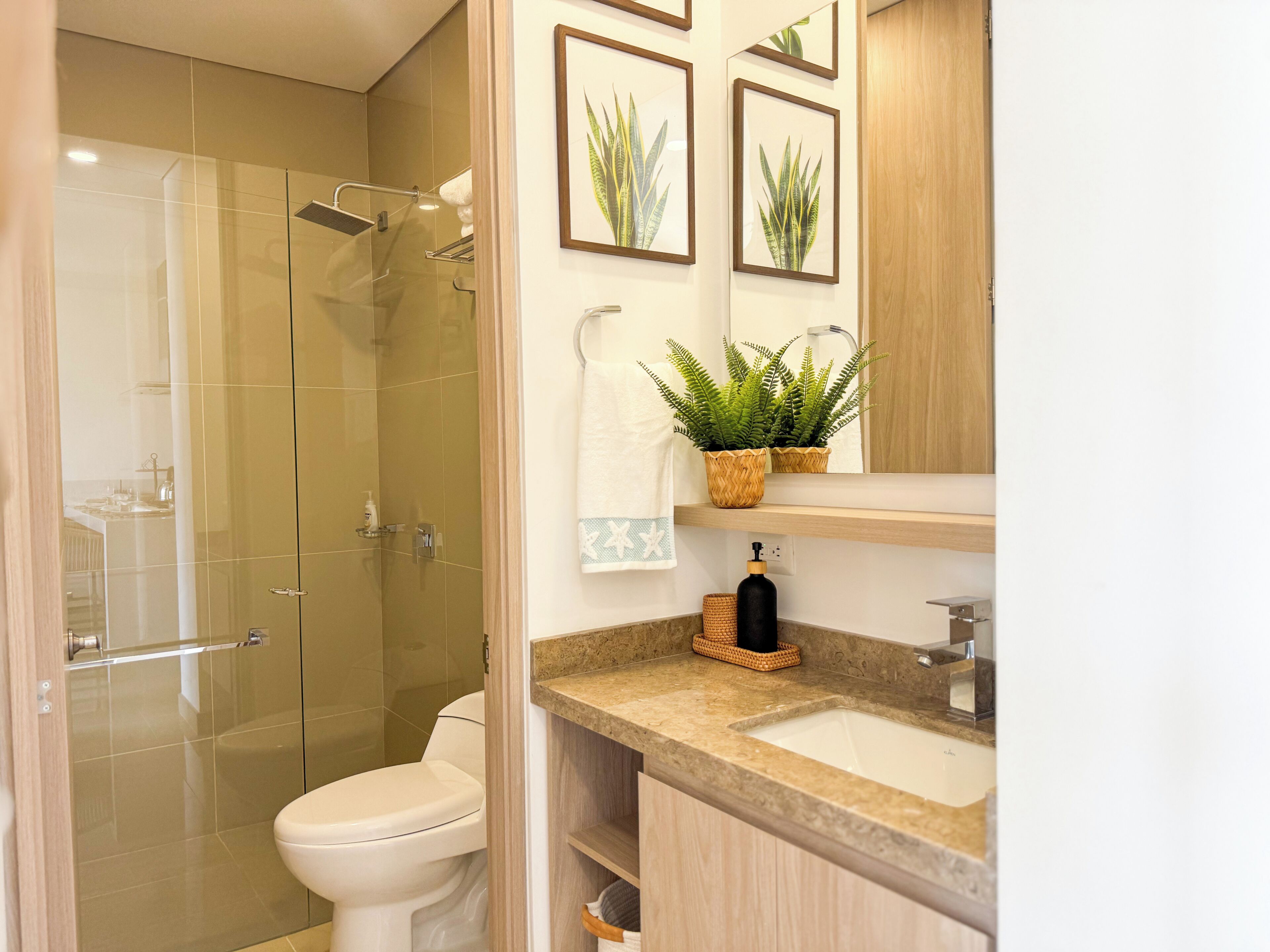 Une salle de bain avec une douche à parois de verre, des toilettes, un lavabo avec un miroir et des tableaux représentant des plantes, accrochés au mur.