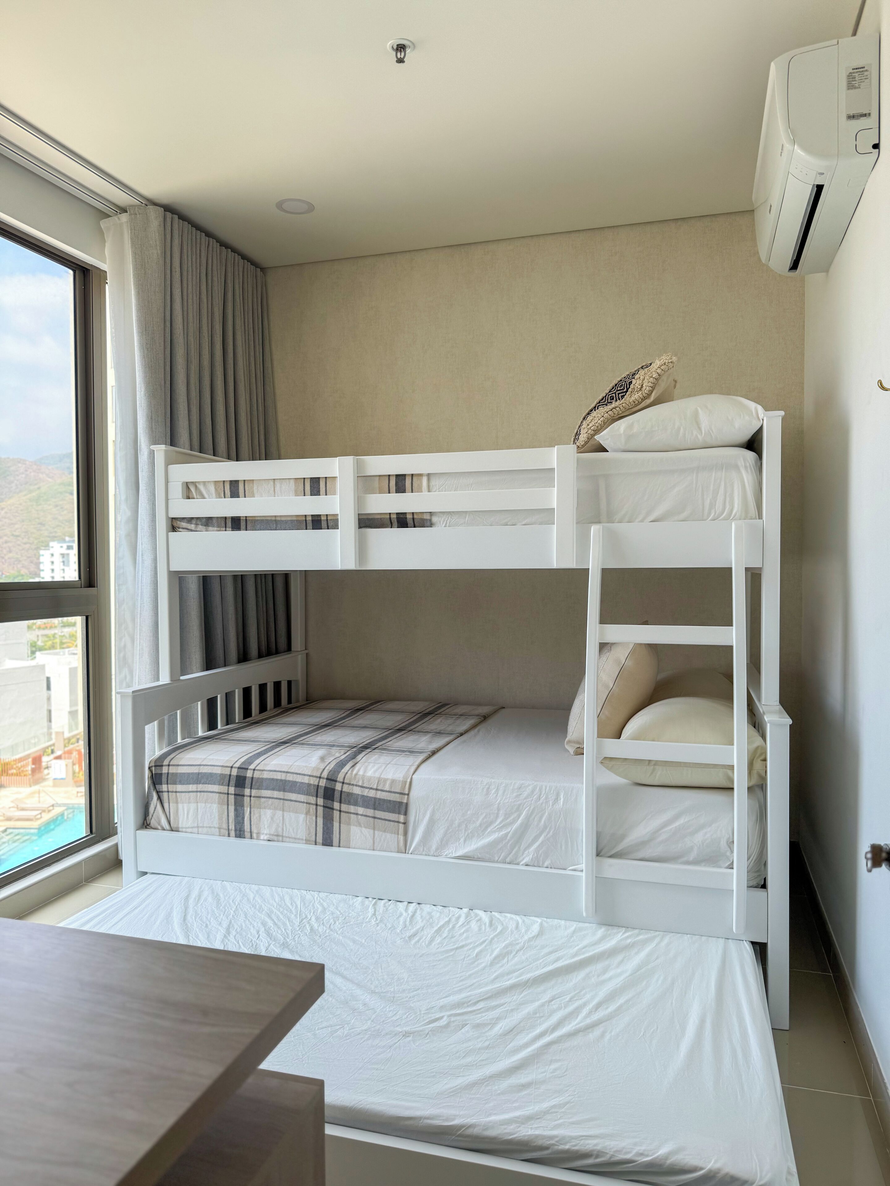 Una cama litera con una colcha a cuadros, una ventana con cortinas y un equipo de aire acondicionado en la pared.