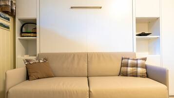 En beige soffa med två kuddar, en vit garderob med ett handtag och hyllor med diverse föremål på vänster sida.