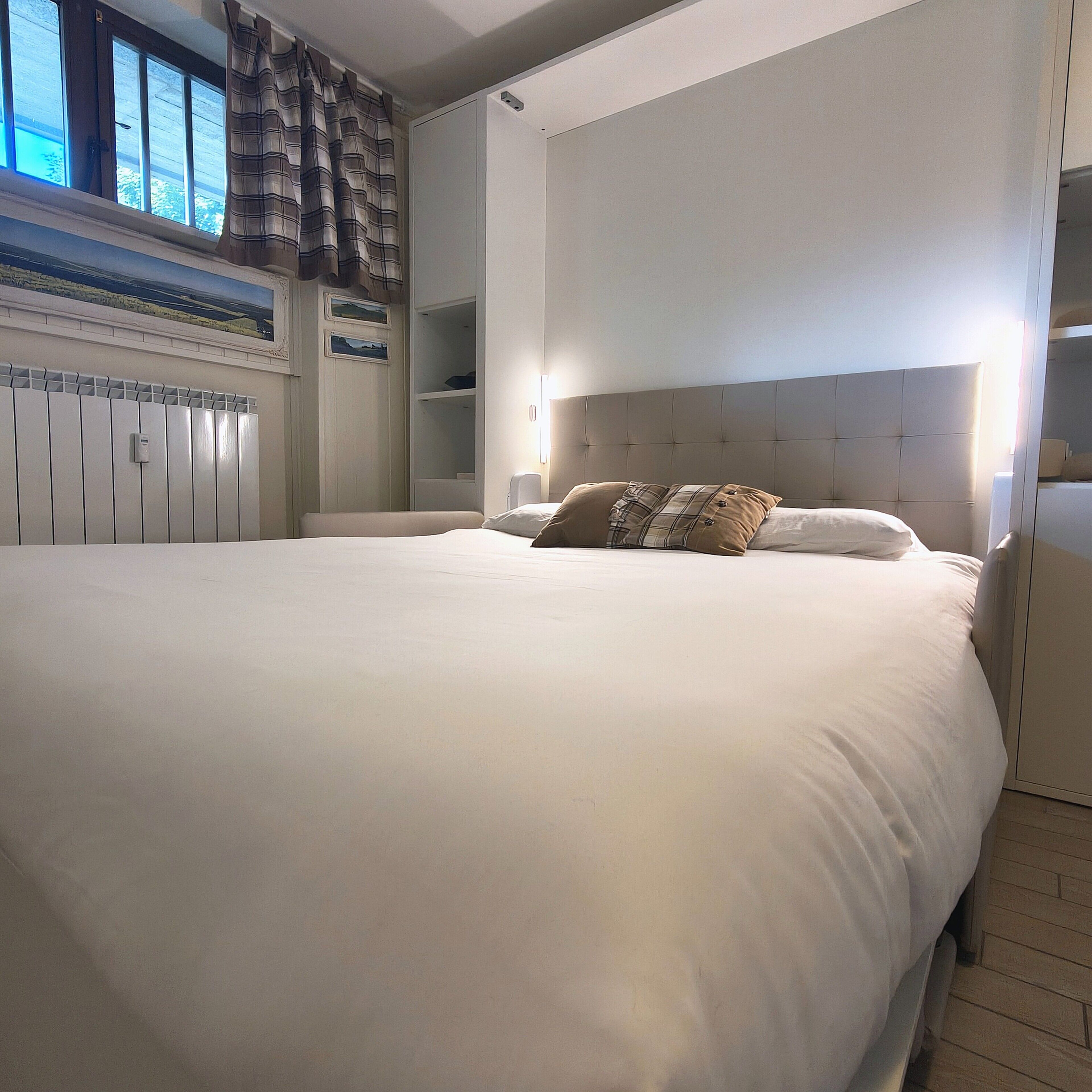 Une chambre à coucher avec un grand lit, une tête de lit, un radiateur et une fenêtre avec des rideaux.
