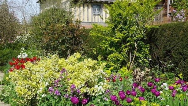 Ein traditionelles Haus mit geneigtem Dach und Schornsteinen, umgeben von einem Garten mit verschiedenen Blumen und Pflanzen.