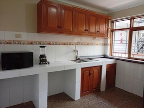 A kitchen with white cabinets, a black microwave, a blender, and a window with a view of a building. - Hostal Cusco de mis Sueños (Cusco)