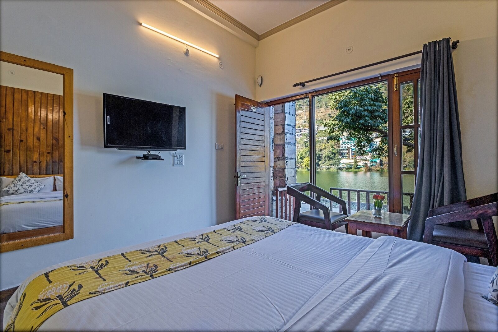 A bedroom with a bed, a TV mounted on the wall, a window with a view of a lake, and a balcony with a chair.
