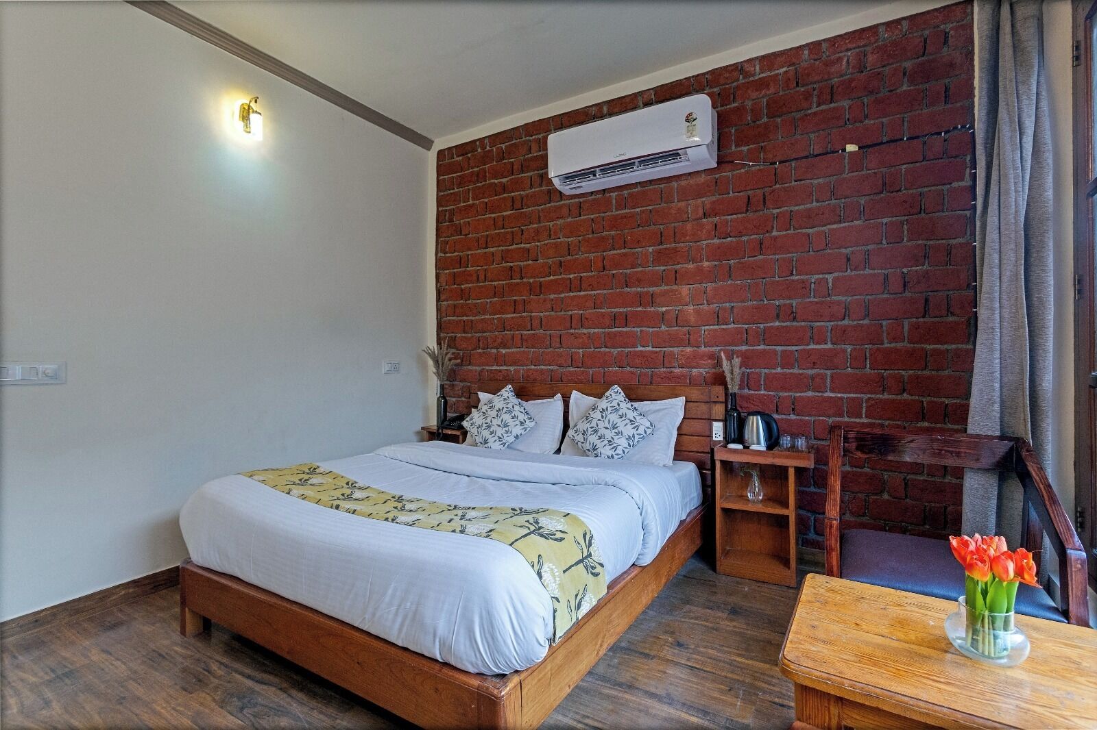 A bedroom with a brick wall, a wooden bed, a nightstand, a table with a vase of flowers, and an air conditioning unit.
