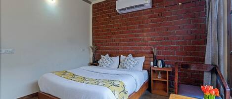 A bedroom with a brick wall, a wooden bed, a nightstand, a table with a vase of flowers, and an air conditioning unit.