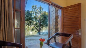 A room with a wooden table and chairs, a window with a view of a tree and a body of water, and a door with a number plate. - Dimmys By the Lake Bhimtal (Bhimtal)