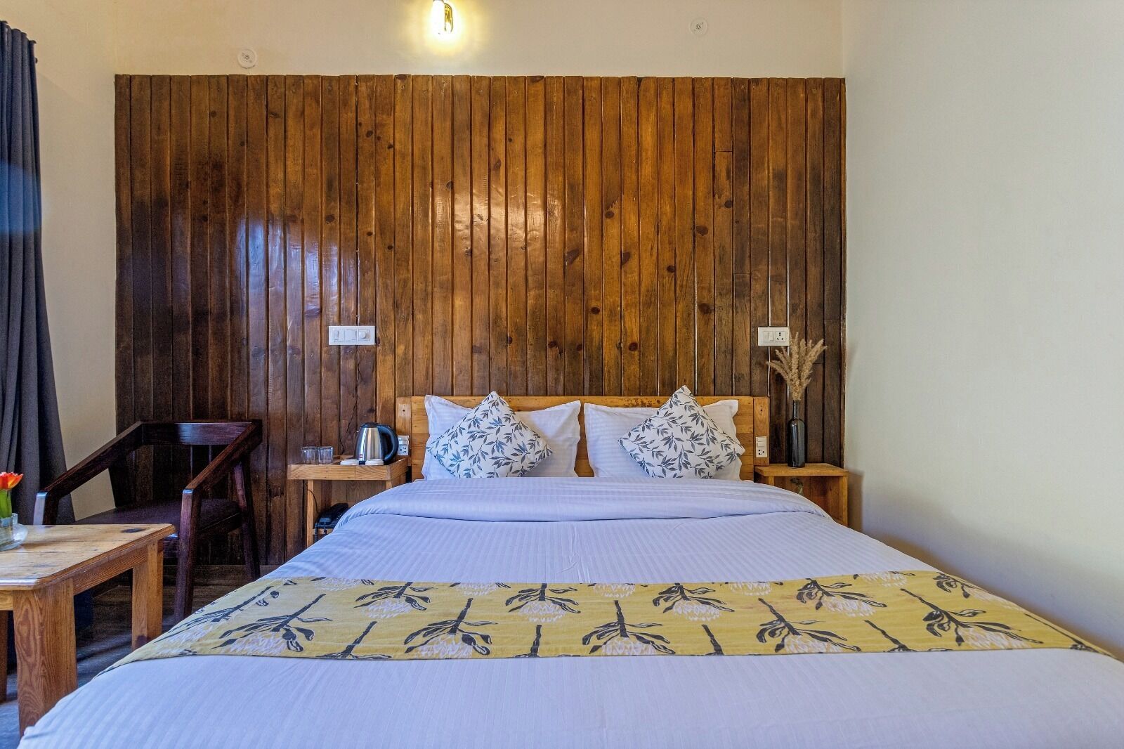 A room with a wooden panel wall, a bed with white bedding and patterned pillows, a wooden nightstand with a kettle, a chair, and a table with a vase.
