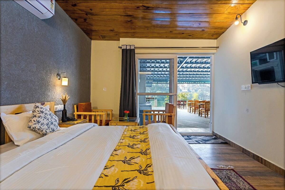 Habitació d'hotel amb dos llits, una taula petita amb un ram, una banca de fusta i vista a la zona de seients exterior a través d'una porta de vidre.
