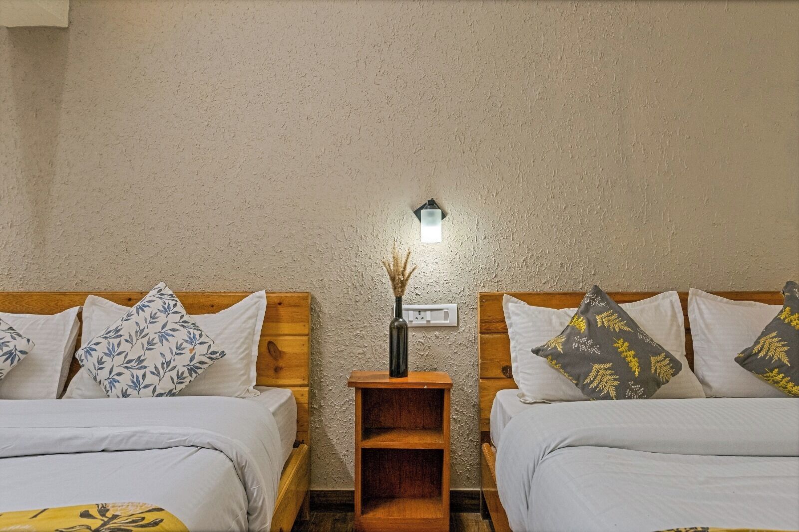Une chambre avec deux lits en bois, chacun doté d’un matelas blanc et d’oreillers à motifs. Une petite table de chevet en bois, sur laquelle se trouve un vase contenant des plantes séchées, est placée entre les deux lits. Une lampe fixée au mur éclaire la pièce.