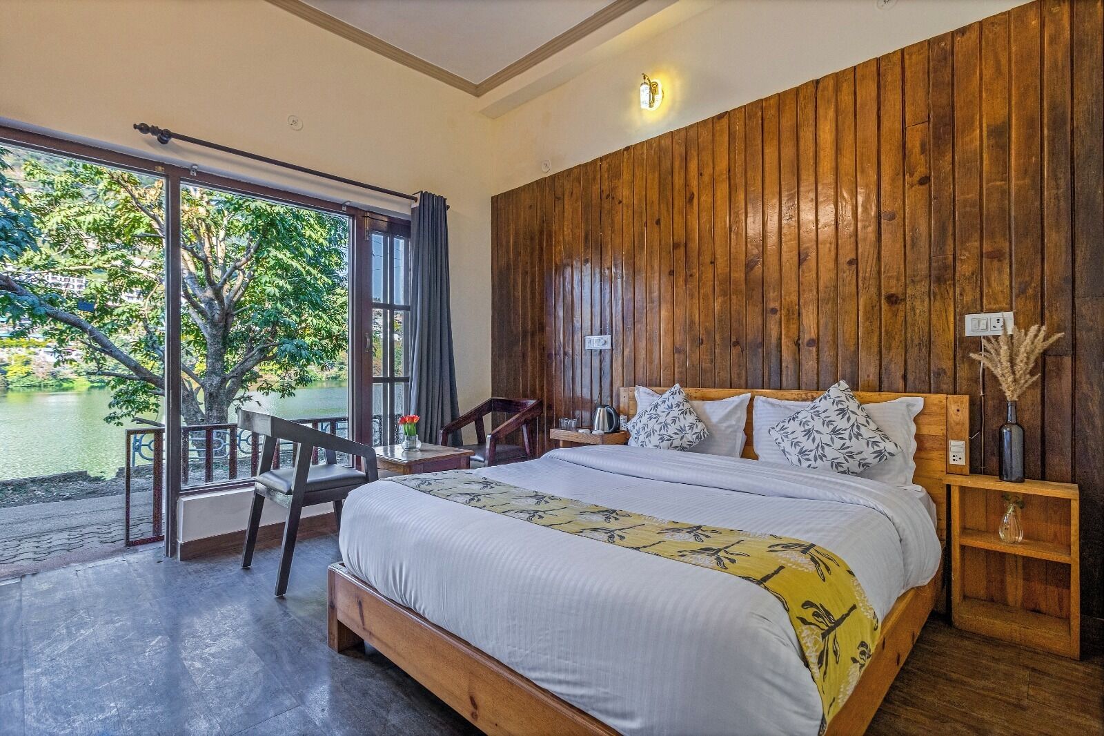 A bedroom with a large bed, wooden paneling on the wall, a nightstand, and a view of a lake through a sliding door.