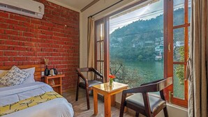 A room with a brick wall, a bed, a table with chairs, and a large window with a view of a lake and hills. - Dimmys By the Lake Bhimtal (Bhimtal)
