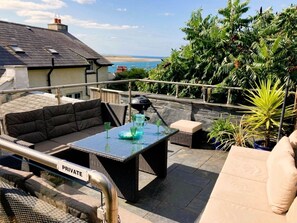 Eine private Terrasse im Freien mit einer Couch, einem Tisch und Stühlen mit Blick auf einen Strand, umgeben von viel Grün.