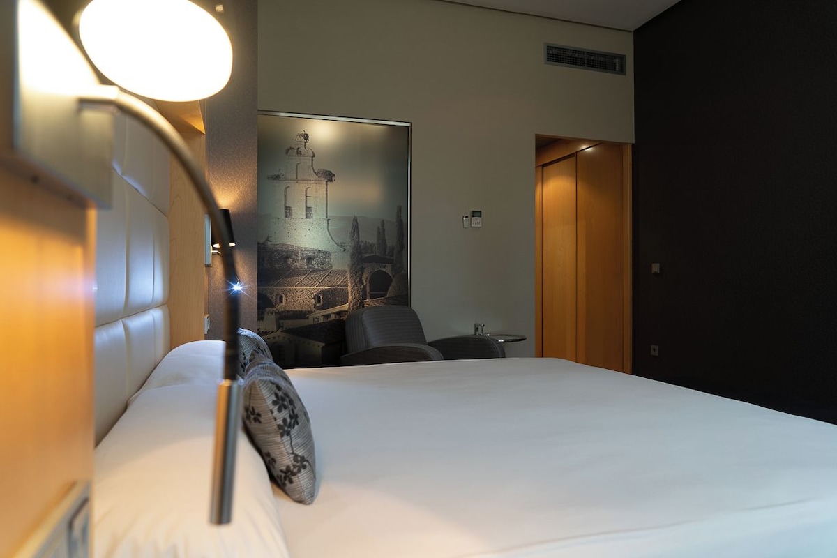 Une chambre d’hôtel avec un lit, un bureau, une chaise, une lampe fixée au mur et une porte.