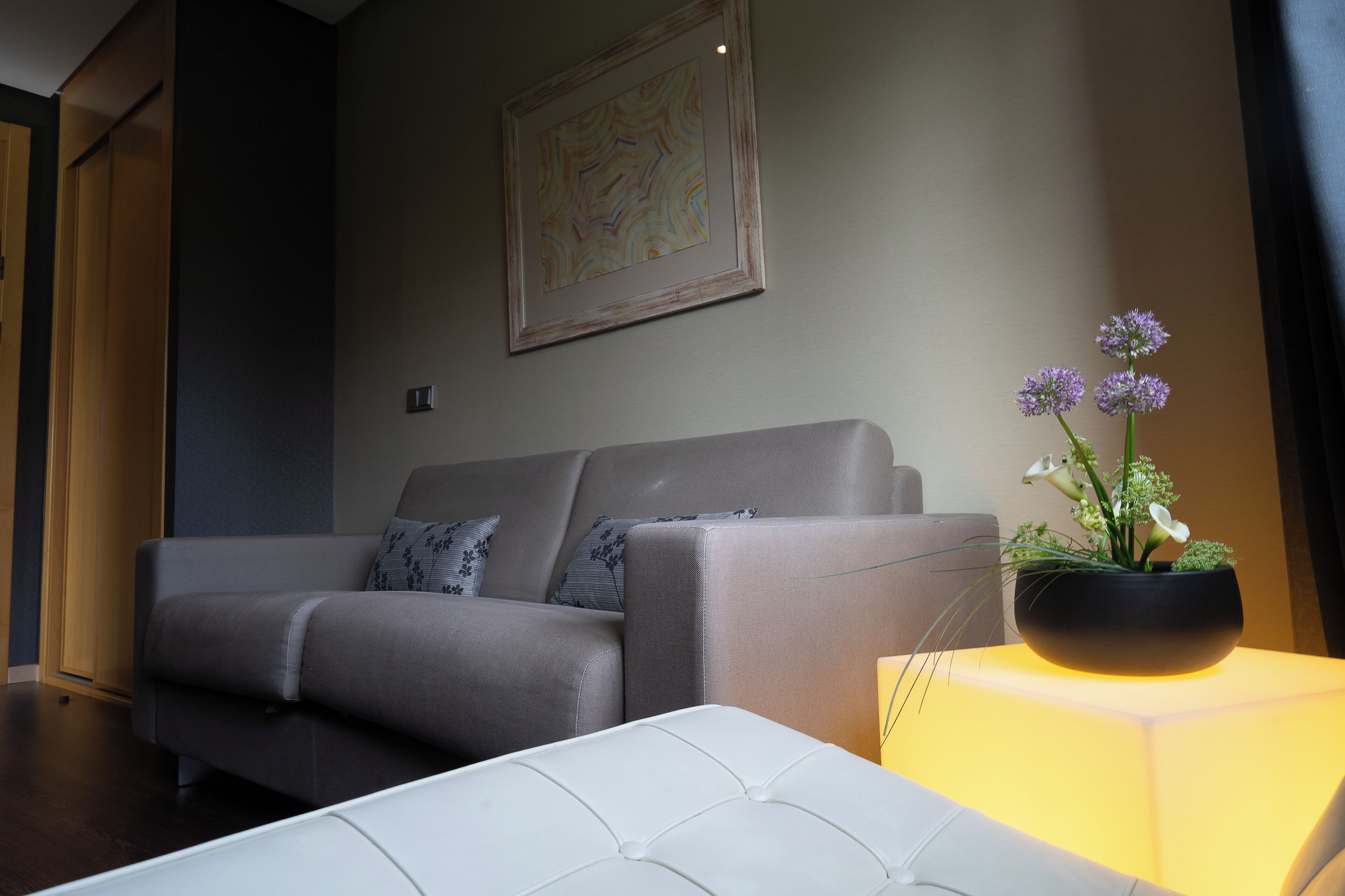 A modern living room with a grey sofa, a white ottoman, and a black vase with purple flowers on a table.