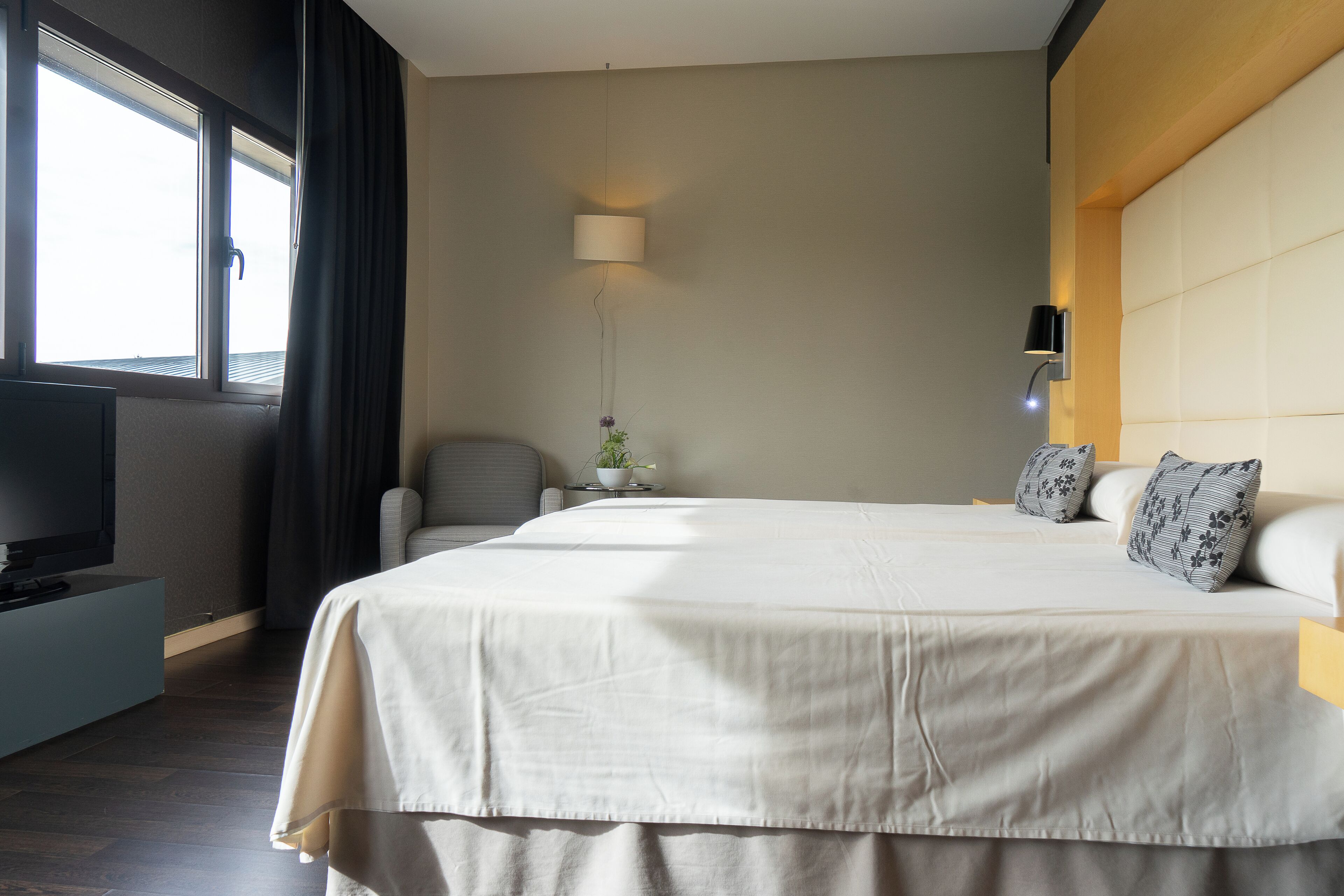 A hotel room with a large bed, a television on a stand, a desk with a lamp, and a window with curtains.