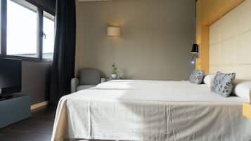 Habitación de hotel con una cama grande, un televisor sobre un mueble bajo, un escritorio con lámpara y una ventana con cortinas.