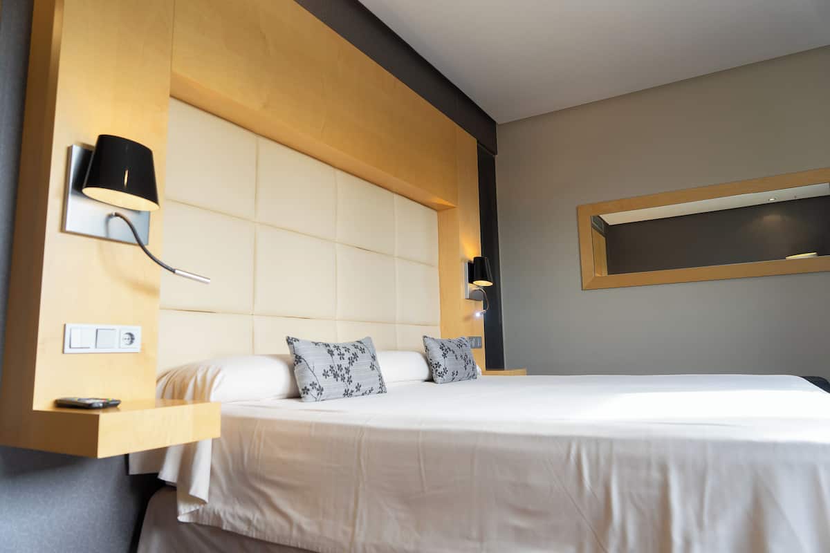 Una habitación de hotel moderna con una cama grande, un cabecero de madera, una mesita de noche con lámpara y una estantería empotrada.