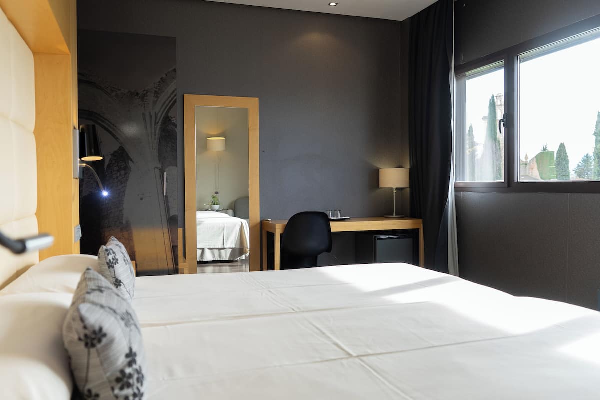 Habitación de hotel con cama, escritorio, silla, lámpara y ventana con vista a árboles.