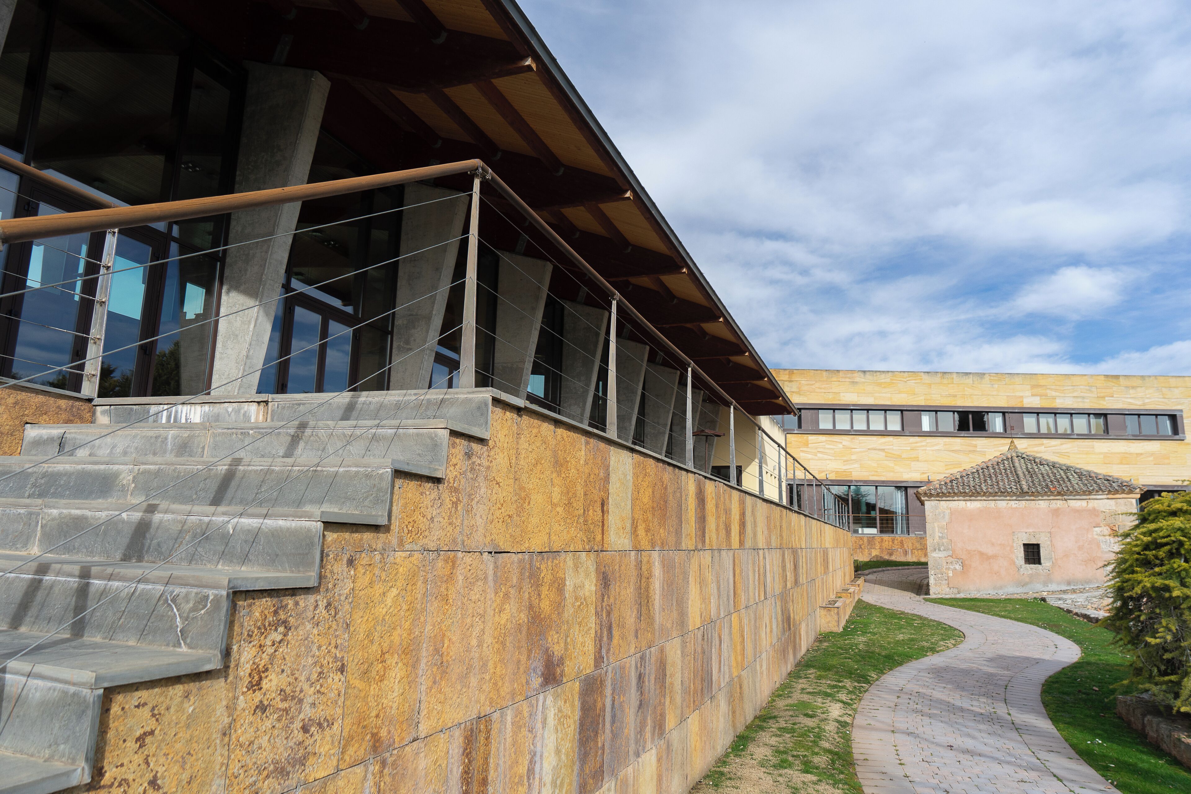 A modern building with a stone staircase leading to a glass entrance, a paved pathway, and a small adjacent structure.