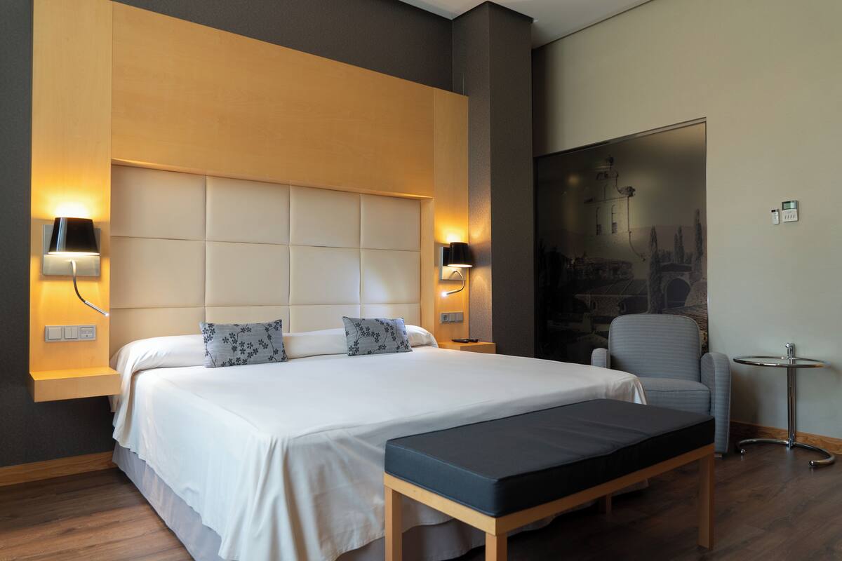 Una habitación de hotel moderna con una cama grande, dos lámparas de noche, una mesita pequeña y una silla.