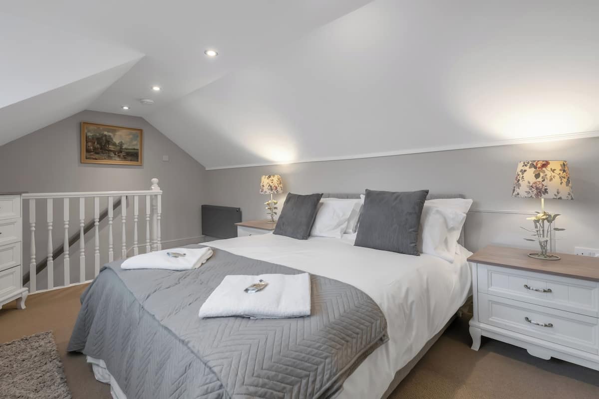 A bedroom with a bed, two bedside tables, a nightstand, a white dresser, a staircase, and a painting on the wall.