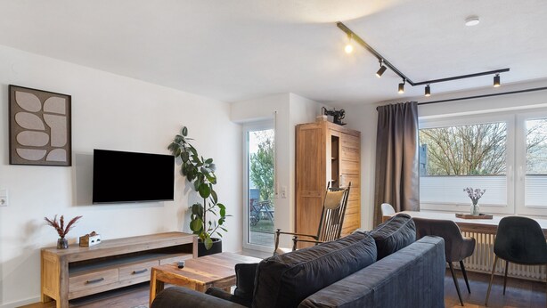 En moderne stue med et fladskærms-tv, en mørk sofa, et træskab og et spiseområde med bord og stole.