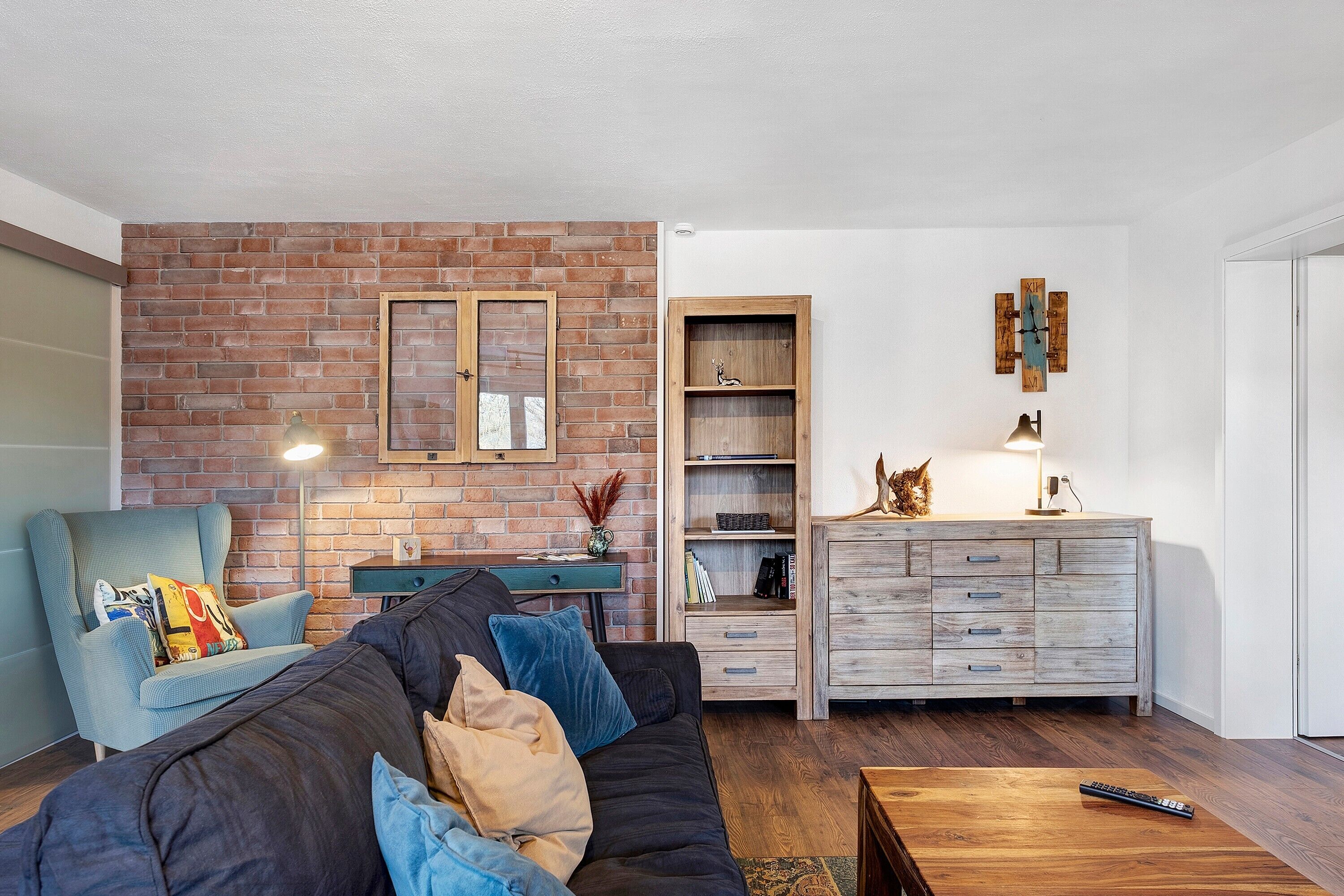 Ein Wohnzimmer mit Backsteinwand, einer Couch mit Kissen, einem Holztisch, einem Bücherregal und einem Schrank.