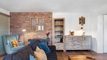 Ein Wohnzimmer mit Backsteinwand, einer Couch mit Kissen, einem Holztisch, einem Bücherregal und einem Schrank.