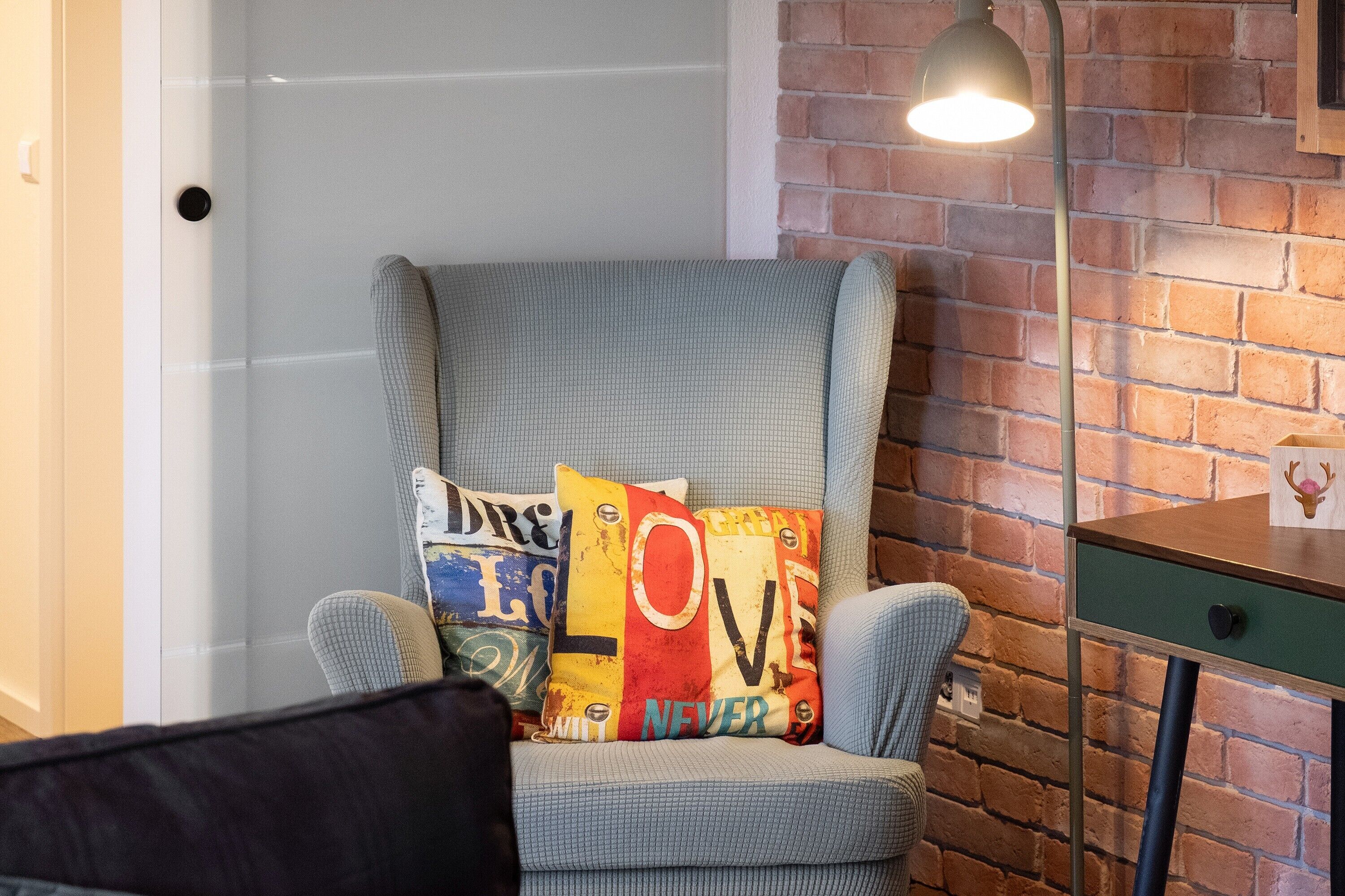 A corner room with a grey armchair, a small table, and a brick wall.
