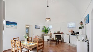 A spacious dining area with wooden chairs and a table, a kitchen with white cabinets, a small refrigerator, and a hanging light fixture.